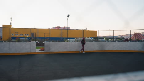 female jogger runs along curved court under twilight sky, viewed through chain link fence with pastel horizon light and urban backdrop of yellow industrial building and high rise apartments at dusk