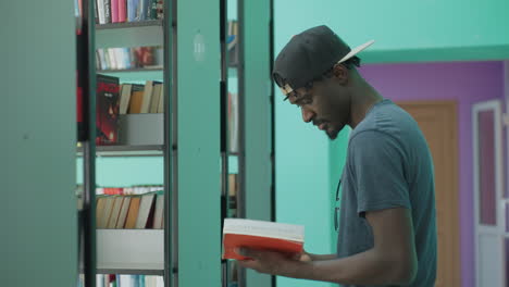 student in cap returns brown book to shelf while skimming red cover volume inside bright library corridor, glancing toward camera, relaxed and focused