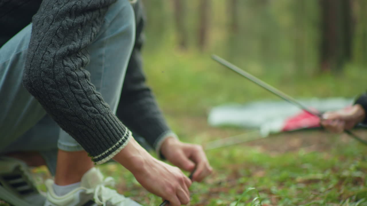 Tent maker fixing pole for camping while partial view of person handing over additional poles is visible, surrounded by green forest environment