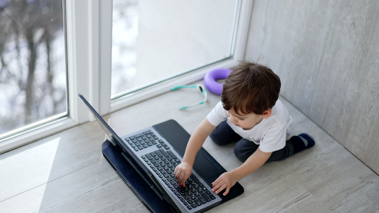 A little boy sits on the floor and plays on a laptop. The child is sitting on the floor near the window and looks at the screen.