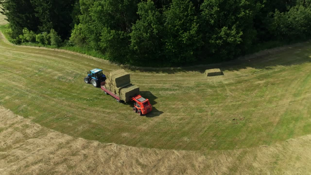 Drone view of a vehicle loading a bale of hay onto a tractor. Harvesting dry grass from a meadow in summer. Czech Republic