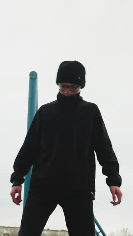 a young boy dressed in a black outfit and glasses carefully walks on an elevated structure, while another boy in a gray sweater hangs on an iron pole in the background