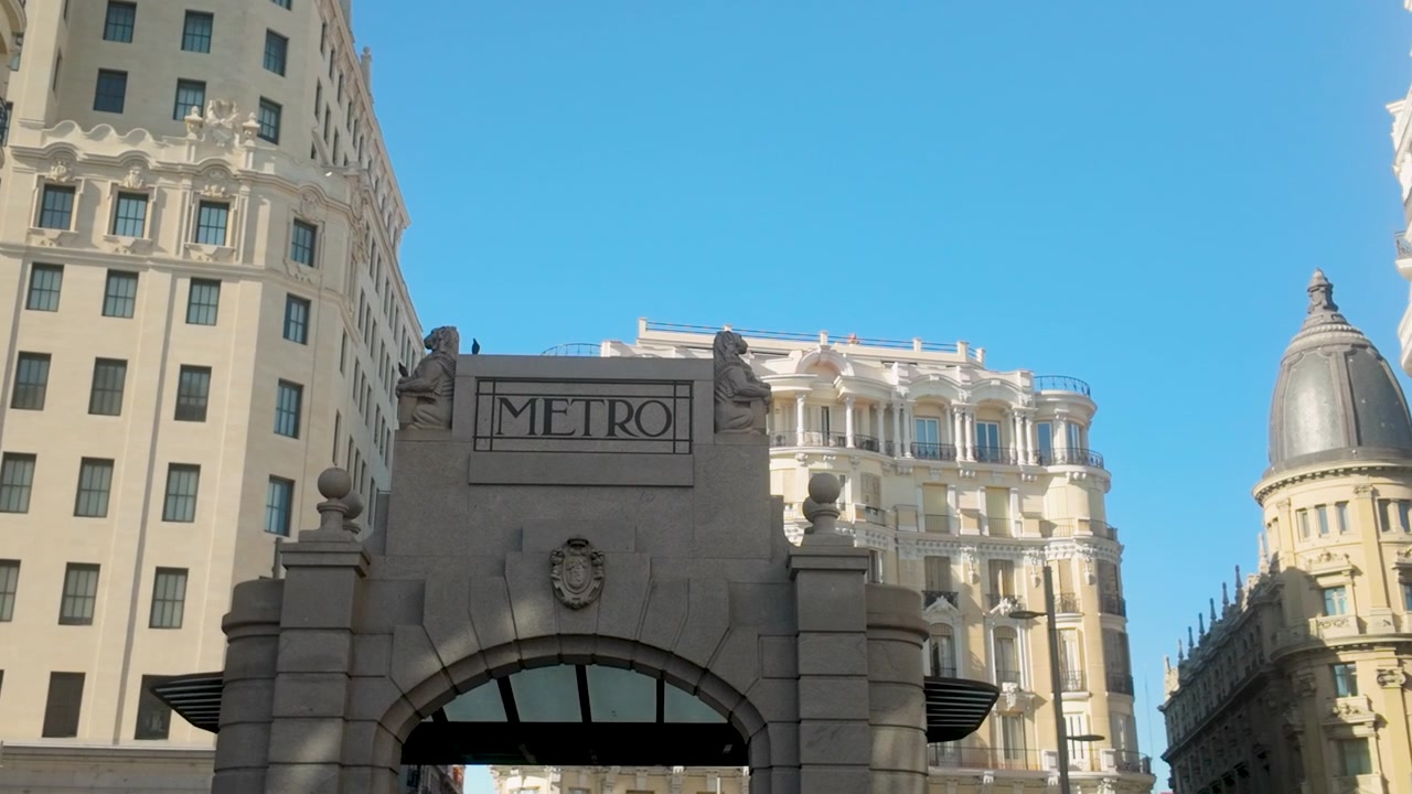 Una toma contrapicada de la icónica entrada de piedra del Metro en Madrid, España, rodeada de grandes edificios históricos bajo un cielo azul claro