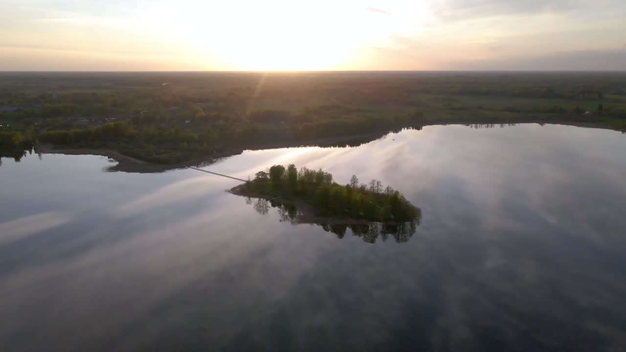 Stunning aerial drone footage circling a small forested island on a calm lake at sunset. Serene reflections on the water and peaceful evening light create a tranquil and scenic atmosphere.