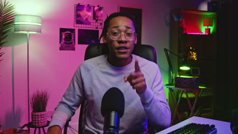 a man talking into a microphone while sitting at a desk with a computer and a headset. he is smiling and appears to be recording a video or podcast.