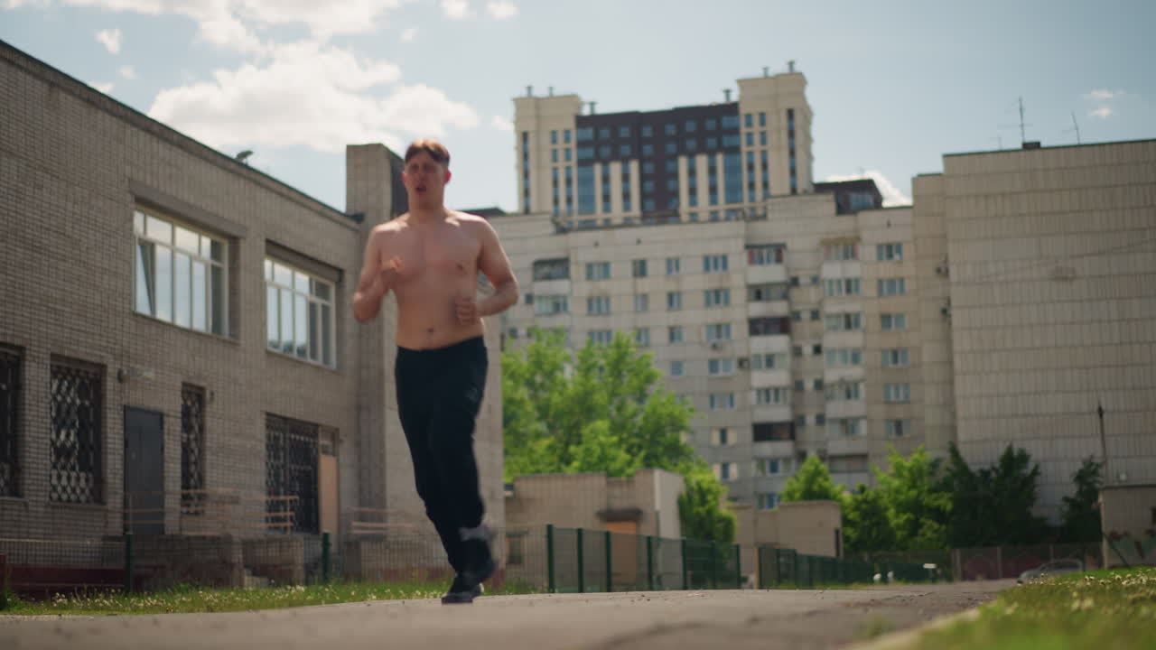 카메라를 향해 마당을 가로질러 달려오는 상의를 탈의한 백인 남성, 힘찬 발걸음과 집중된 시선, 햇빛이 비치는 건물과 포장도로, 활기찬 훈련 의지와 도시적 결단력