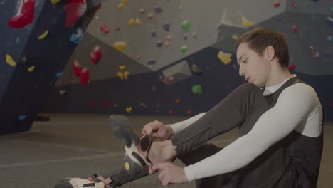 young male athlete sitting in bouldering gym and putting climbing shoes on 1