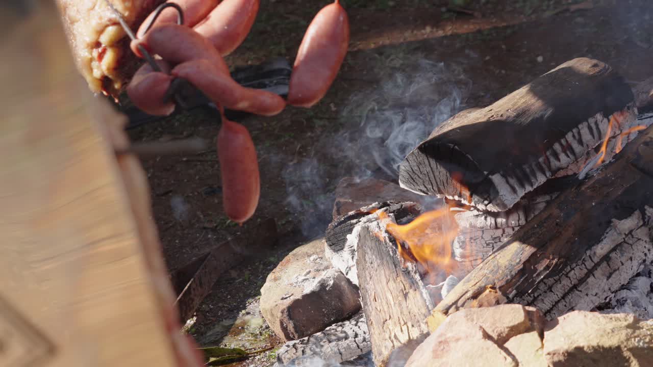 Argentine chorizos hang over a wood fire as they begin to cook, captured in slow motion to show smoke and glowing embers rising from the rustic pit during an outdoor asado in natural sunlight
