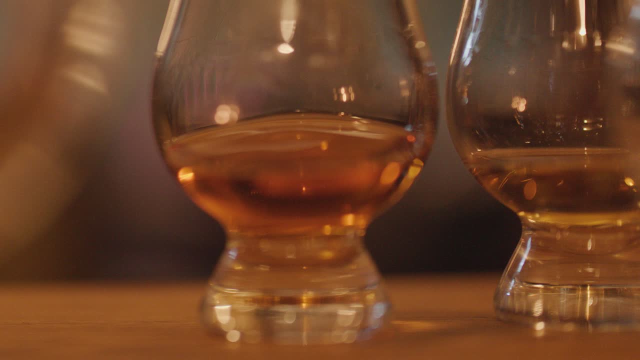 A hand sets whisky glasses on a wooden bar, warm candle-like lighting creates a cozy, intimate atmosphere with shallow depth of field and soft background blur