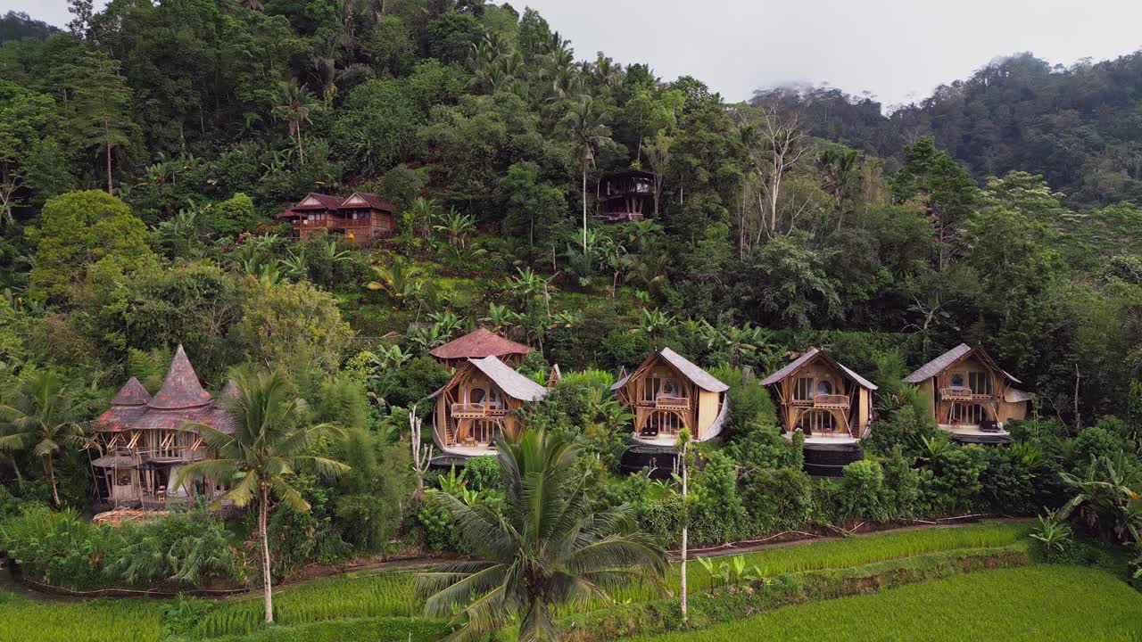 Aerial drone view capturing stunning bamboo villas nestled in a tropical mountain valley surrounded by lush rice terraces in Bali, Indonesia, highlighting peaceful eco-living and natural harmony