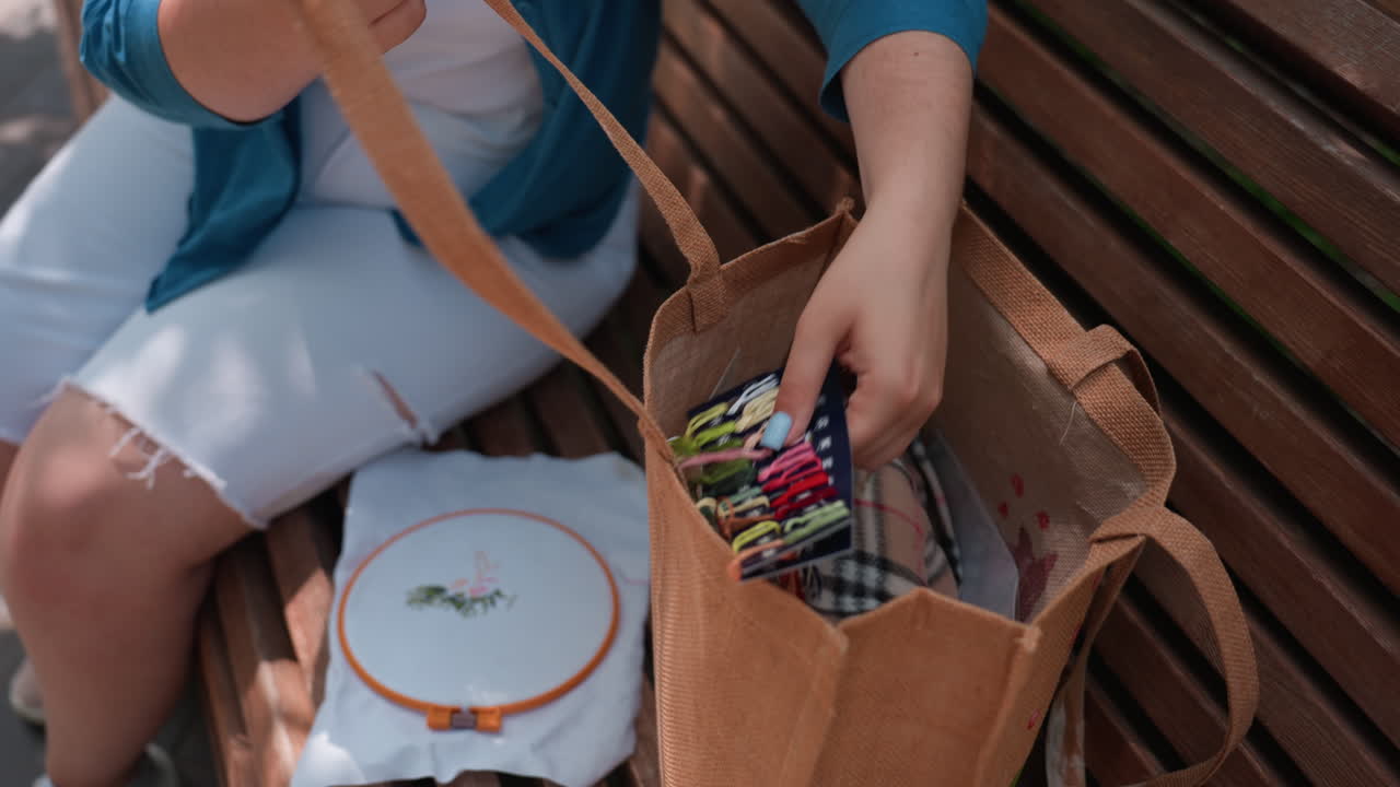 Close up of girl sitting on wooden bench returning colorful embroidery materials into brown tote bag beside her, sunlight illuminating fabric and threads, calm atmosphere of creativity