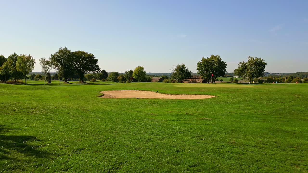 Low-altitude drone shot showing a sand bunker, golf flag, trimmed green grass, and surrounding trees under clear sky
