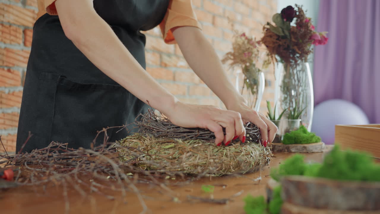 Female florist in apron crafting rustic wreath using twigs and moss on wooden table with plants and flowers in glass vases, emphasizing handmade decoration process, creativity, seasonal decor design