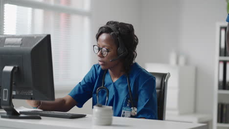 mujer negra médico usar bata blanca gafas y auriculares hablando con el cliente uso de videollamada. debido al brote de la pandemia de coronavirus terapeuta trabajando de forma remota proporcionar ayuda a los pacientes de la clínica por videollamada