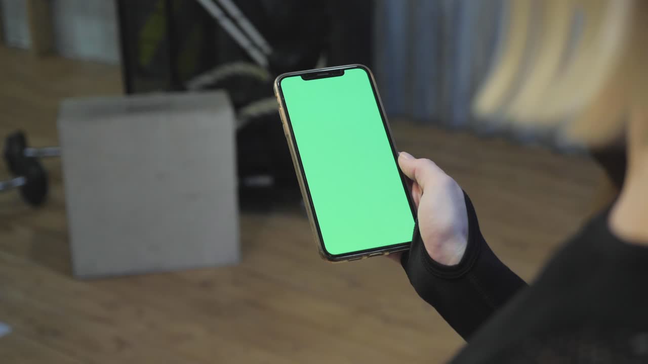 una joven atlética sostiene un teléfono inteligente en la mano, muestra una pantalla chromakey verde, mira una aplicación deportiva, está en el gimnasio