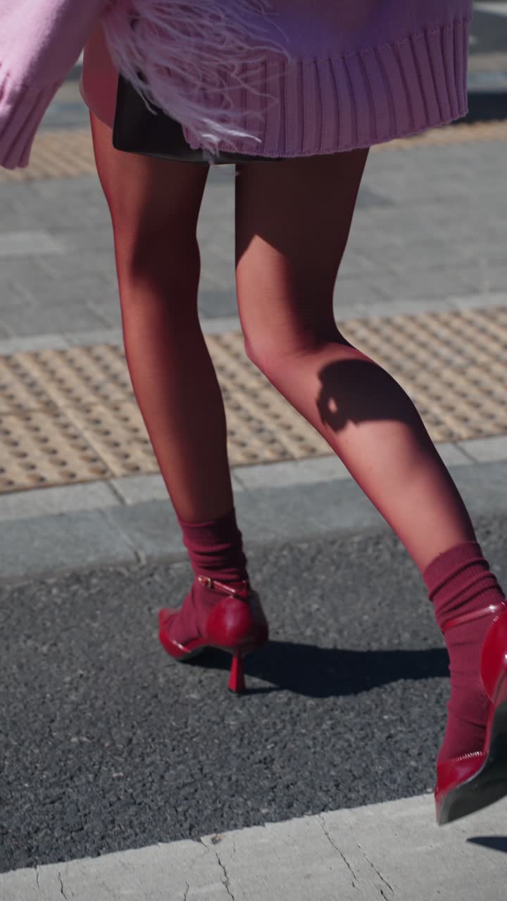 Vertical: A confident woman strides across a crosswalk wearing bold red heels and matching socks, exuding poise and elegance under the bright sunlight in a bustling urban setting - tracking
