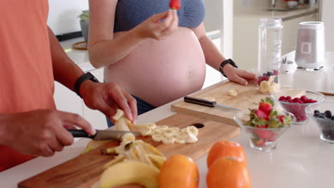 preparing healthy smoothie, pregnant woman holding jar while partner slicing banana