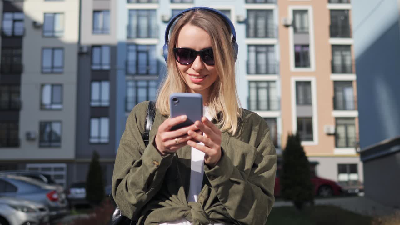 Beautiful blond smiling girl wearing casual clothes and sunglasses choosing music from app playlist. Young woman listening to music on her smartphone in wireless headphones enjoy good quality sound.