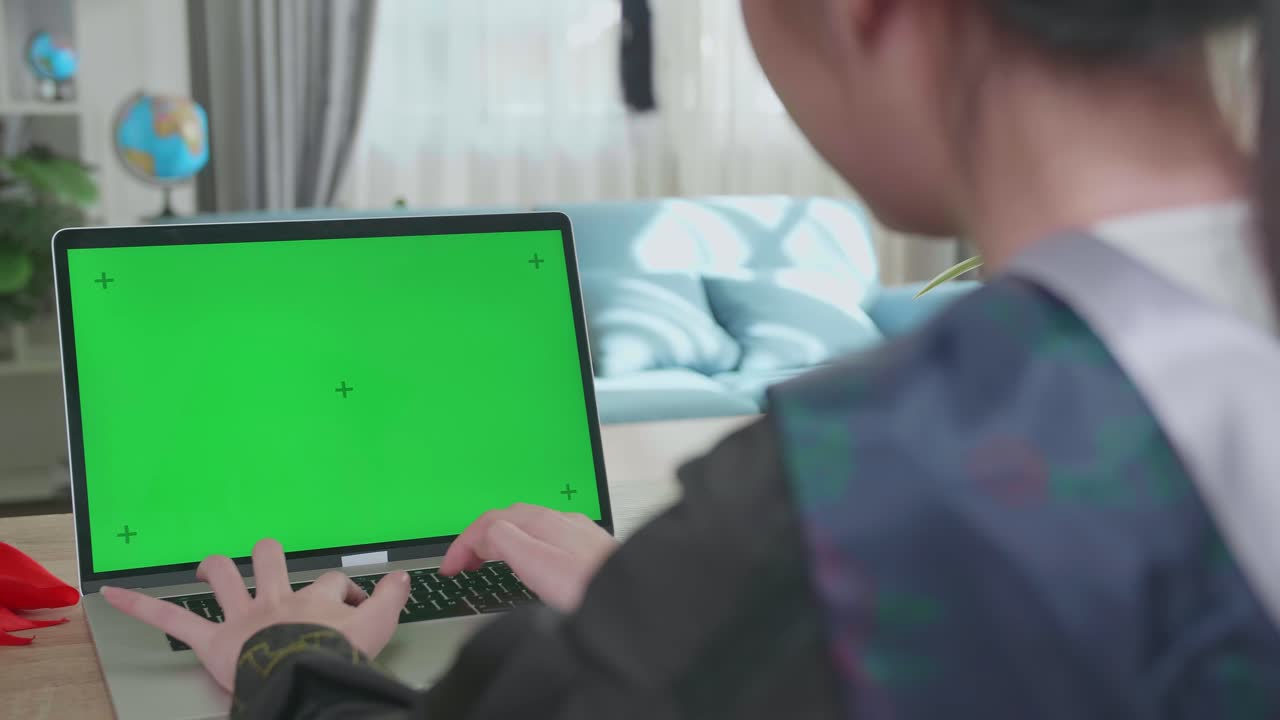 Over The Shoulder Shot Of Asian Woman Wearing A Graduation Gown And Cap Use Laptop Computer With Green Mock-Up Screen In Living Room