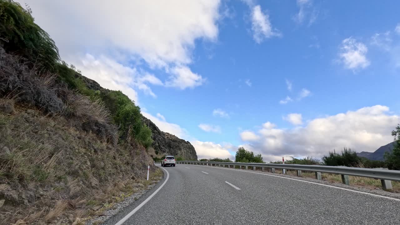 Vehicle drives winding mountain road by lake, daylight, partly cloudy sky, smooth forward camera motion
