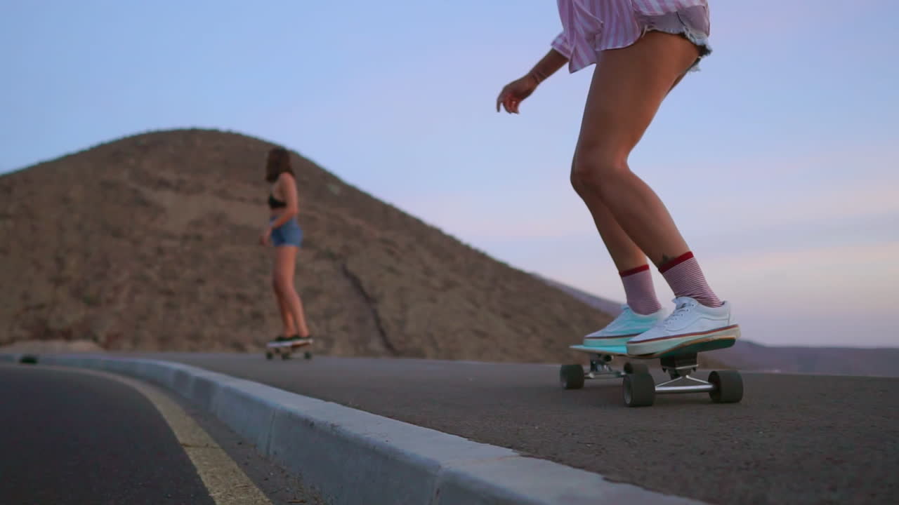 en medio de la belleza de la puesta de sol, dos amigos con pantalones cortos participan en skateboarding en cámara lenta en una carretera, con montañas y un cielo impresionante como telón de fondo