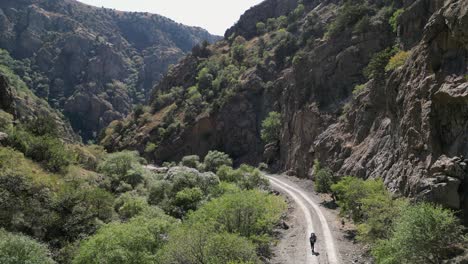 Backpacker-hikes-on-dirt-road-in-rugged-narrow-rock-canyon,-high-angle