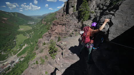 Woman-With-Backpack-on-Climbing-Route-Holding-Rocks,-Moving-Above-Green-Valley,-Slow-Motion