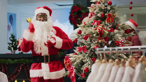 Cheerful-worker-dressed-as-Santa-Claus-ringing-bell-and-setting-xmas-gifts-under-beautifully-decorated-Christmas-tree,-trying-to-attracts-shoppers-to-xmas-adorn-clothing-store