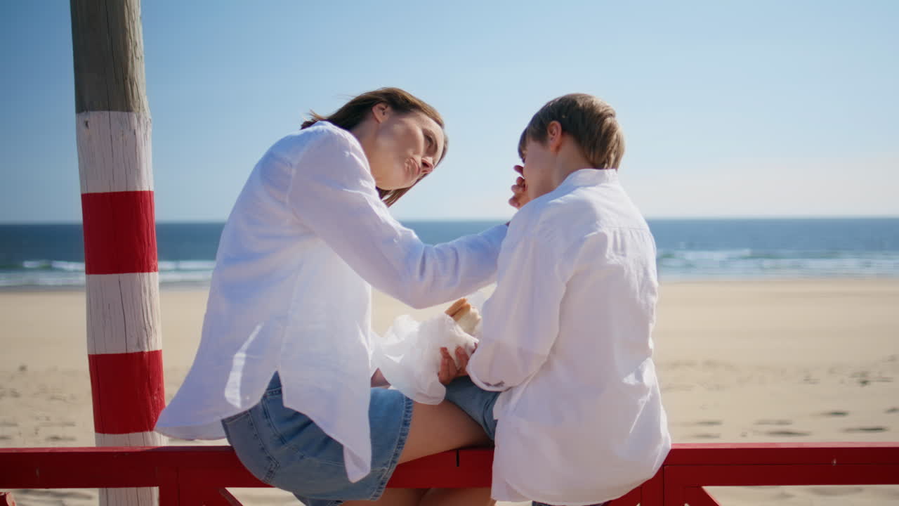 Mother caressing beloved son sitting red fence at sunny beach. Relaxed woman