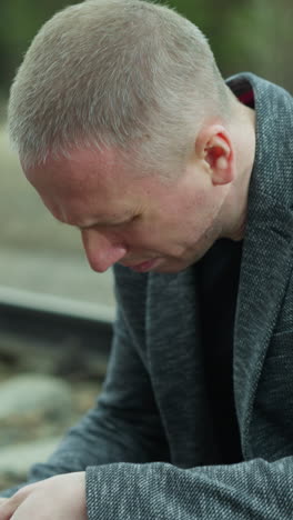 a close view of a man in a gray jacket handling a handgun while sitting down beside a railway track, intently inspecting the firearm, the background shows a blurred view of a green forest