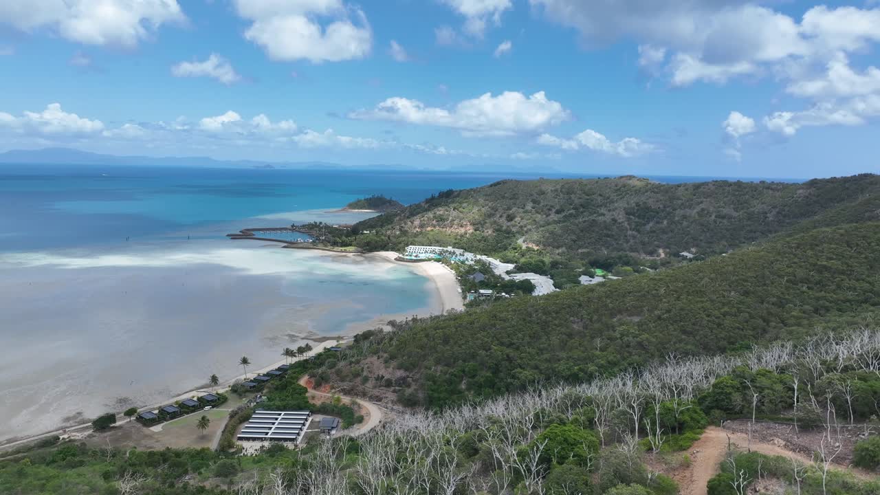 Sakte dronesporingsopptak på dagtid, beveger seg sidelengs langs fjellsiden for å avsløre Hayman Islands buede strand, grunt buktlandskap og turkise Whitsundays-farvann, Queensland, Australia