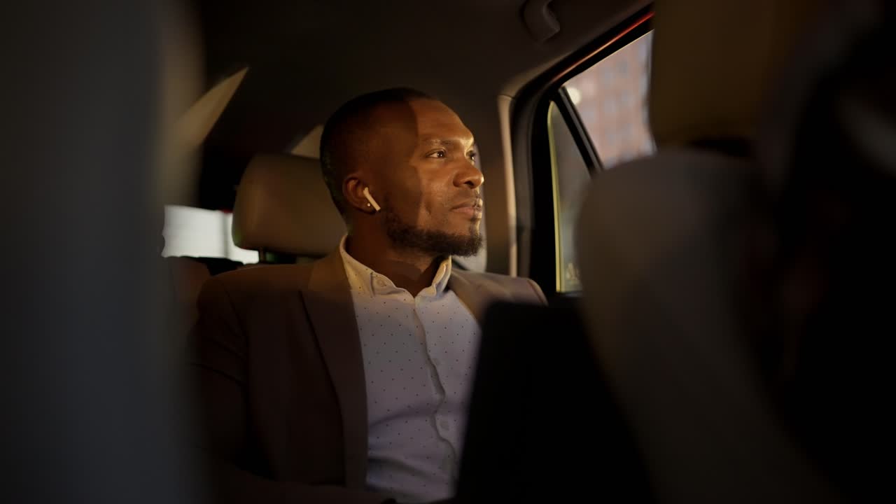 갈색 슈트를 입은 검은 피부를 가진 자신감있는 남자는 무선 헤드폰으로 음악을 듣고 현대 자동차 내부에서 그의 비즈니스 여행 중에 은 저녁에 창밖을 바라보고 있습니다.