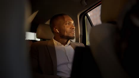 a confident man with black skin in a brown suit listens to music on wireless headphones and looks out the window at a sunny evening during his business trip in the interior of a modern car