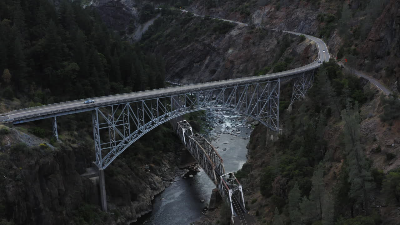 Drone view flying over an extremely tall metal bridge spanning a small river in a deep mountain valley.