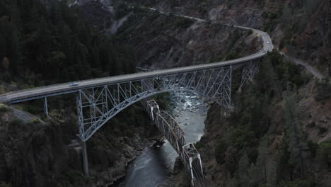 drone view flying over an extremely tall metal bridge spanning a small river in a deep mountain valley.