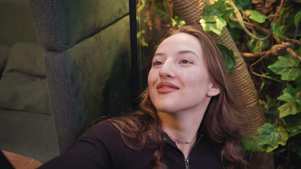 Woman with wavy hair smiles brightly while taking selfie near vibrant indoor plant. Surrounded by green leaves and warm lighting, her cheerful expression highlights connection with nature