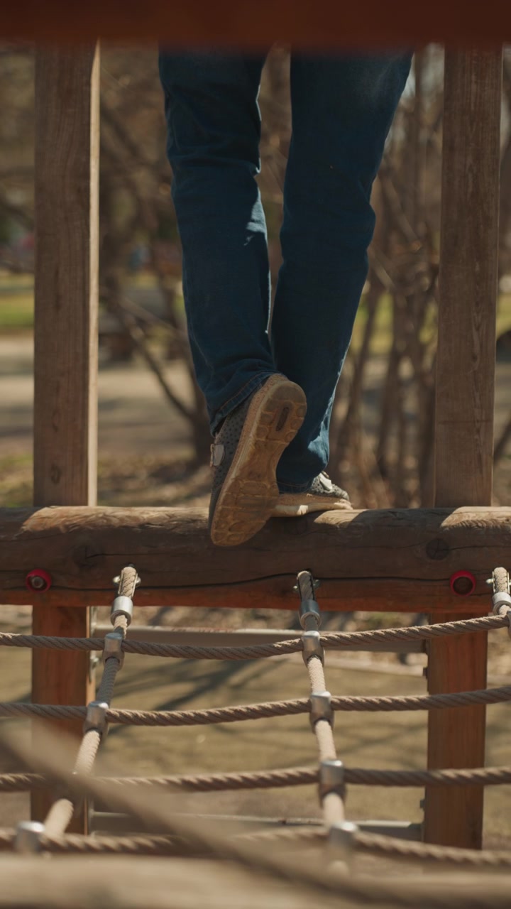 Primer plano de las piernas y pies de una persona caminando cuidadosamente por una pista de obstáculos de madera al aire libre con cuerdas. El enfoque se mantiene en el movimiento a través de cada tablón, resaltando el equilibrio en acción. Luz natural suave.