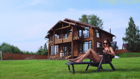 Una-Hermosa-Mujer-Esbelta-Yace-En-Una-Tumbona-En-Traje-De-Baño-Con-Una-Tableta-En-El-Fondo-De-Una-Gran-Casa-De-Campo-Con-Gafas-De-Sol-Y-Trabaja-Remotamente-Durante-Las-Vacaciones