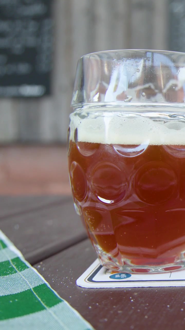 Una persona solleva un boccale di birra in vetro da un tavolo di legno alla luce del giorno, poi lo appoggia su un sottobicchiere.