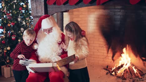 santa is reading a book for a boy and a girl near a fireplace with a fire and a christmas tree