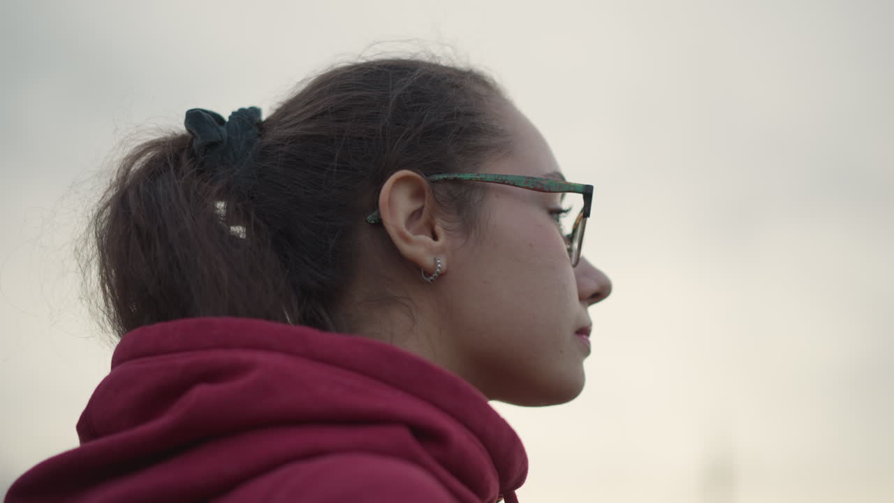 Student Adjusting Hoodie And Looking Upward At Sky CloseUp. Glasses Reflect Soft Clouded Light, Ponytail And Layered Bracelets, Introspective Expression, Urban Dusk Atmosphere And Quiet Determination
