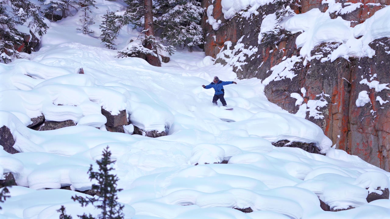 Snowboarder professionista di backcountry sulle Montagne Rocciose del Colorado, che scende da una scogliera a cuscino con un salto aereo, ripreso da drone. Località come Aspen, Snowmass, Vail Pass. Terreno ripido e innevato con campi di massi, in un paesaggio invernale con neve fresca e profonda.