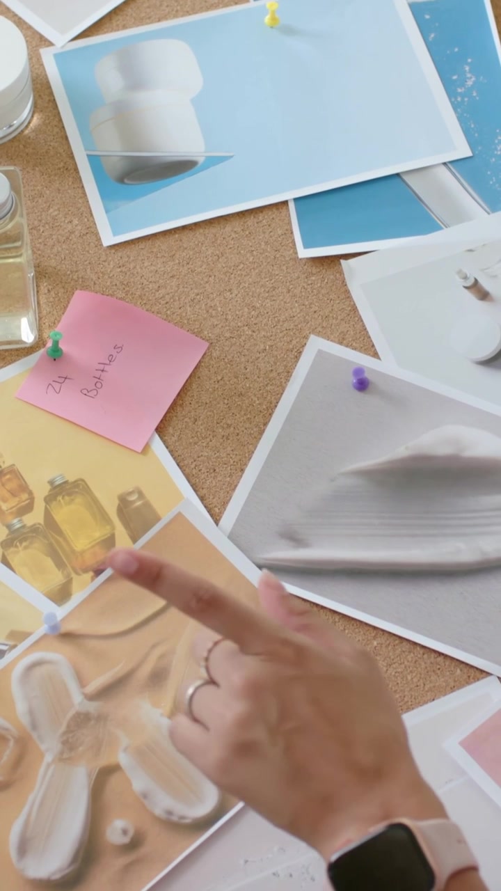 Video vertical: Manos que alisan impresiones en un tablero de corcho para refinar el diseño, con reloj y alfileres.