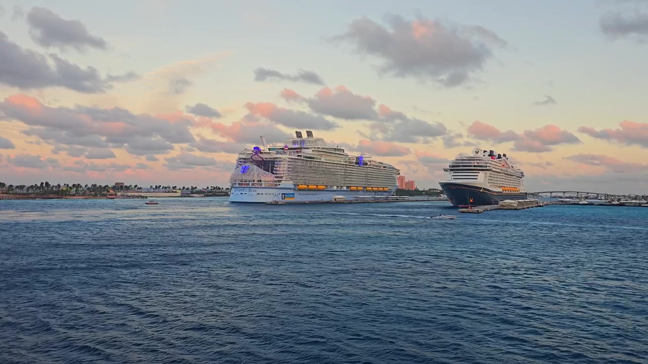 Et bredt luftfoto af to gigantiske krydstogtskibe, herunder Utopia of the Seas, der ligger til kaj i havnen i Nassau, Bahamas. Smuk gylden solnedgangshimmel med bløde lyserøde skyer over blåt vand.