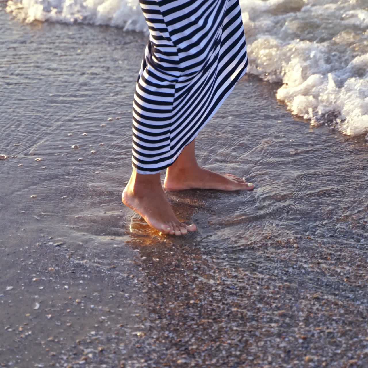 Woman playing with water on beach. Female legs in water. Woman in long dress splashing and playing water by feet on foamy waves background.
