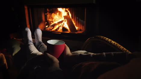 a man warms his feet by the fireplace holds in his hands a red cup with a hot drink