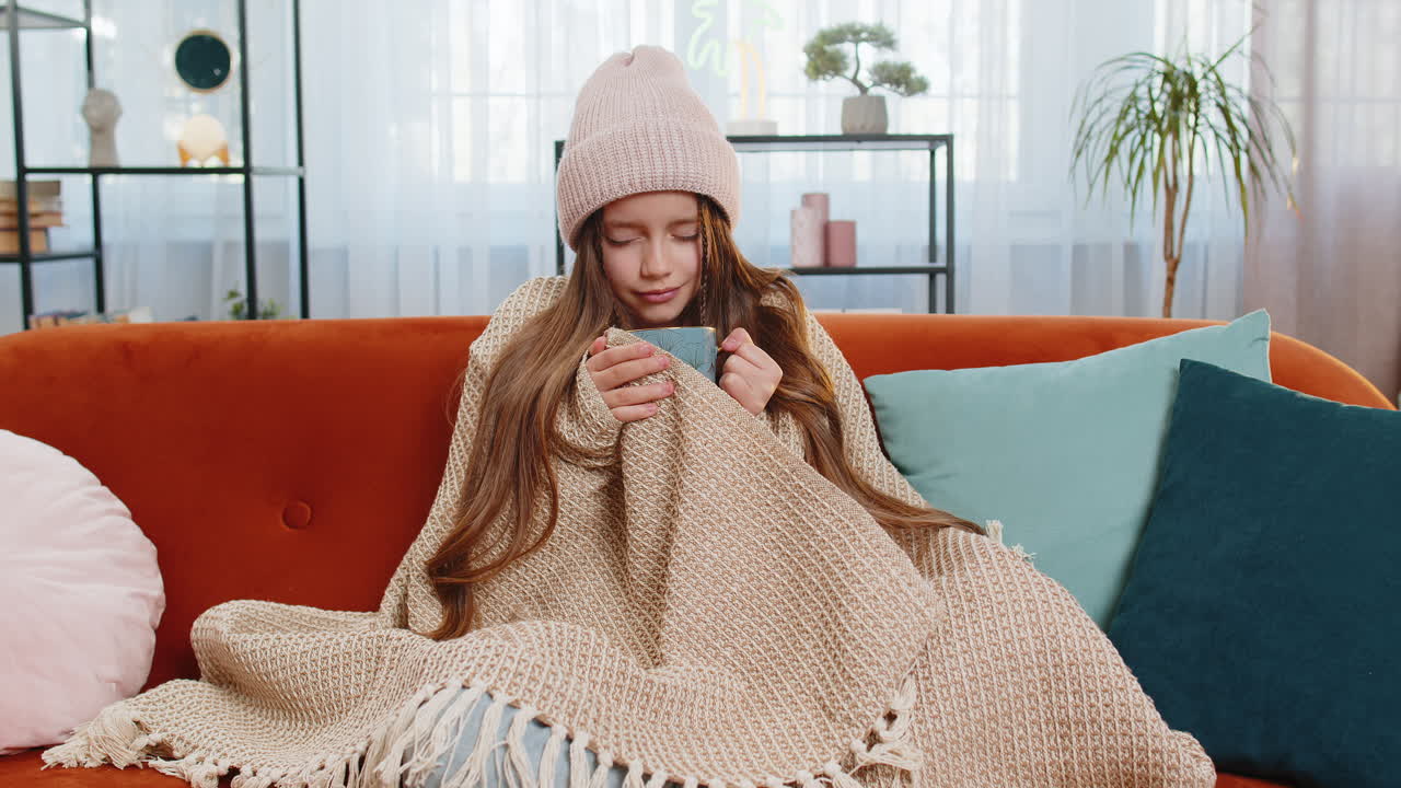 아픈 소녀 아이는 담요로 감싸져 차가운 물에 떨고 집에서 뜨거운 약초 차를 마시고 있습니다.