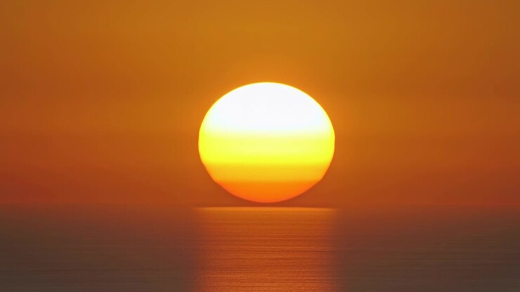 Low sun disk rises above calm sea, shifting upward while narrow vertical reflection lengthens across smooth water, with warm orange sky, soft glow, centered horizon and serene coastal mood