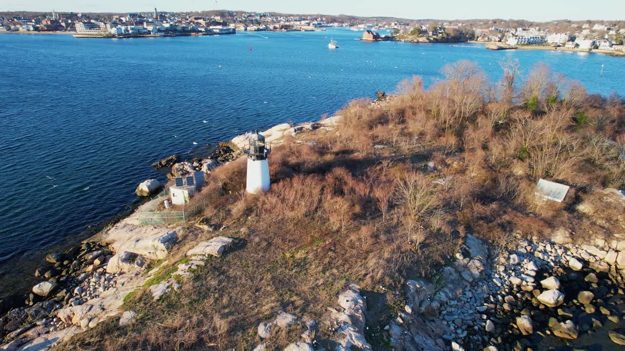 Orbita aerea di un faro a Gloucester, Massachusetts, con costa rocciosa, acqua blu del porto e paesaggio costiero panoramico che crea una classica atmosfera marittima del New England in una giornata di sole