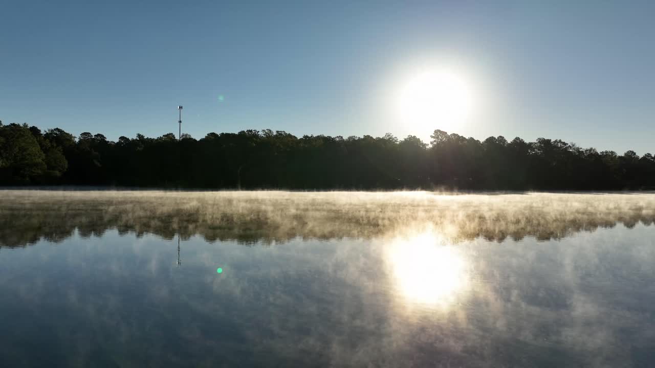 Sunrise light shines through thick steam fog as mist rises above a calm lake surface